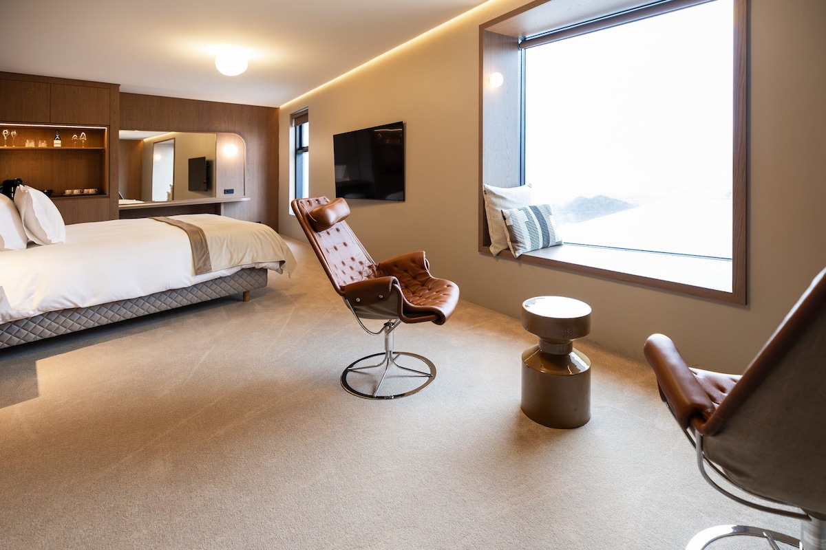 Een moderne hotelkamer met een bed, een zithoek met een fauteuil, een televisie en een raam met uitzicht.