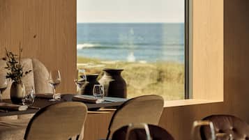 Una mesa de comedor con sillas, un florero con plantas y vista al océano a través de un ventanal grande.