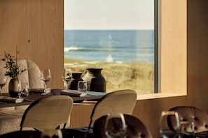 A dining table with chairs, a vase with plants, and a view of the ocean through a large window. - Black Sand Hotel (Sveitarfélagid Ölfus)