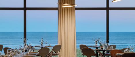 A restaurant with tables set for dining, large windows offering a view of the sea, and a curtain dividing the space.