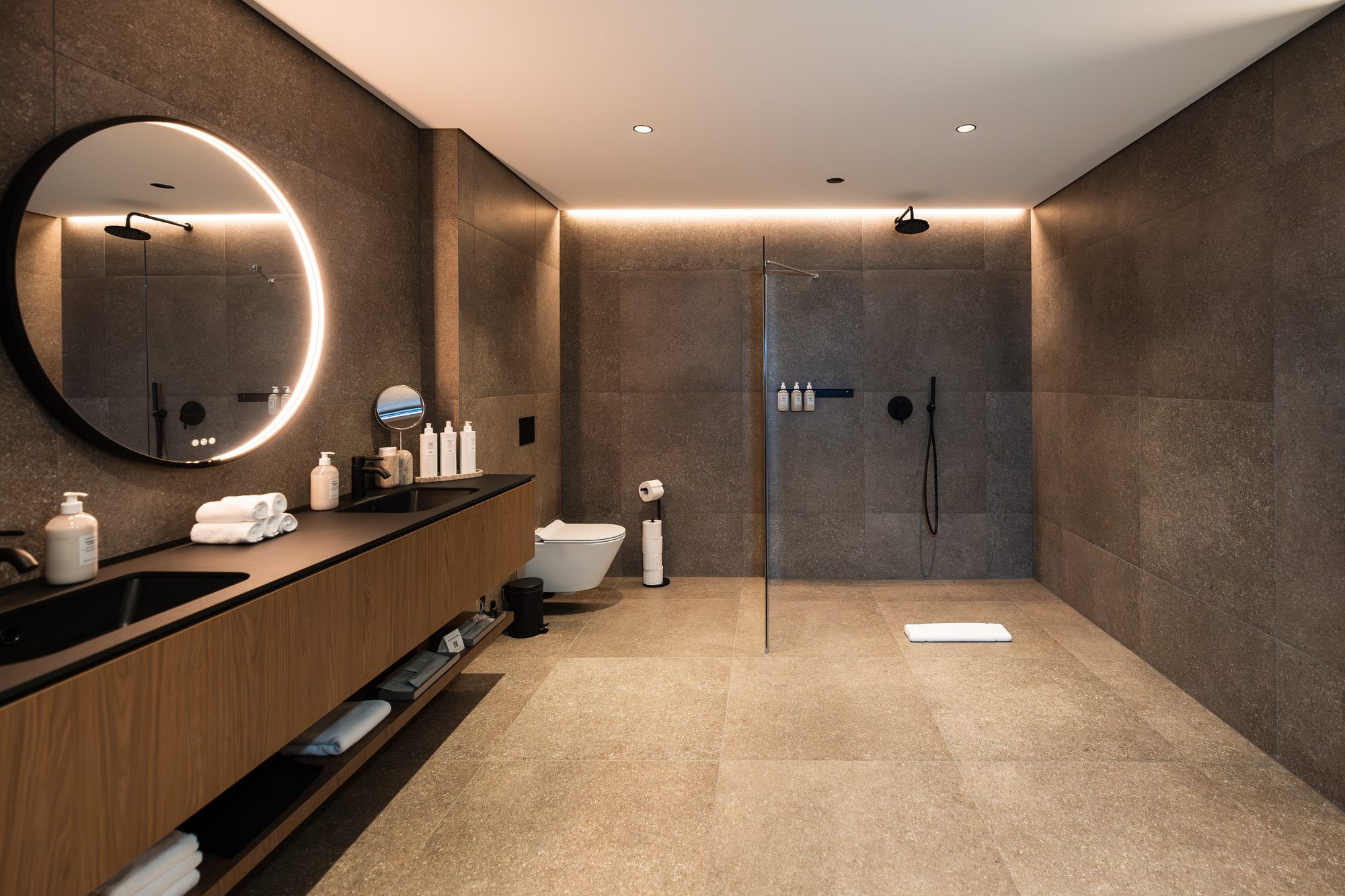 A modern bathroom with a wooden vanity, a large mirror, a round mirror, a shower with a glass door, and a toilet.