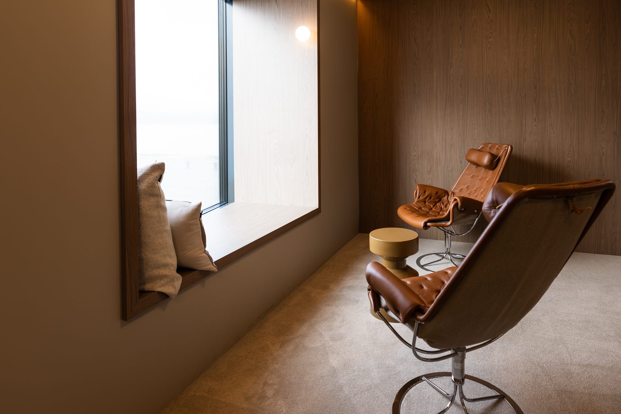 Una habitación con ventana, un sillón de cuero marrón, una mesa redonda y un espejo sujeto a la pared.