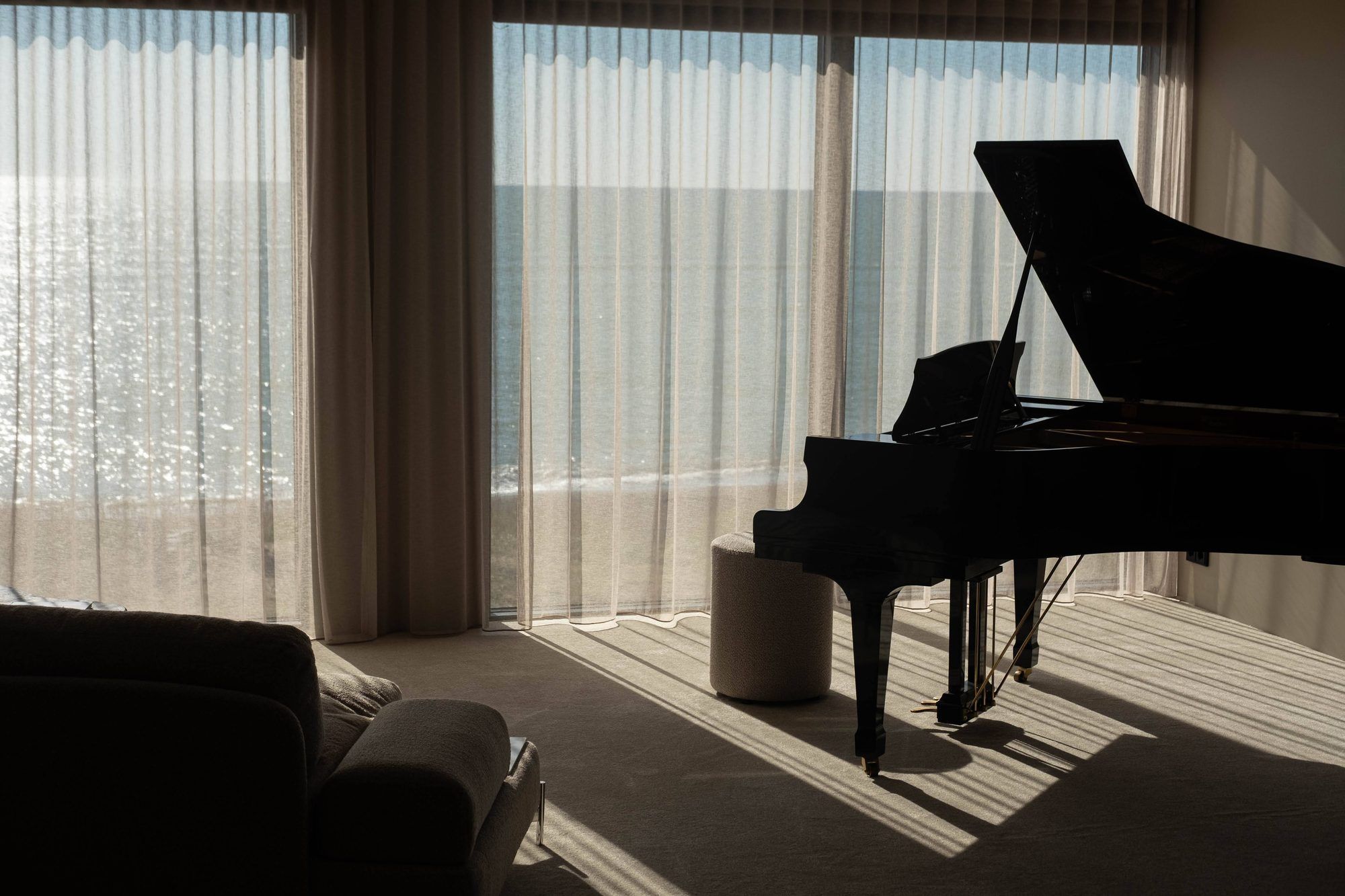 Un piano de cola en una habitación con ventanas amplias, un sofá y un pequeño reposapiés.