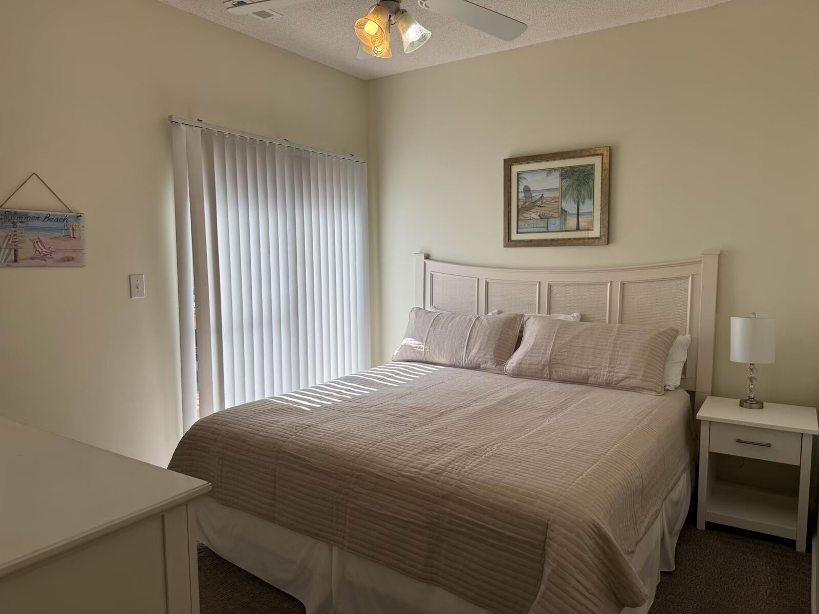 Une chambre bien rangée, avec un grand lit, une table de chevet, une commode, un ventilateur de plafond et un tableau accroché au mur.