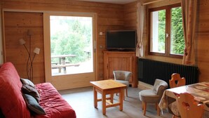 Ein gemütliches Blockhaus-Interieur mit Holzausbau, einer Couch, einem Tisch, Stühlen, einem Fernseher und großen Fenstern mit Blick auf viel Grün.