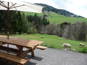 A wooden picnic table with a white umbrella, set against a rural landscape with sheep grazing and houses in the background.