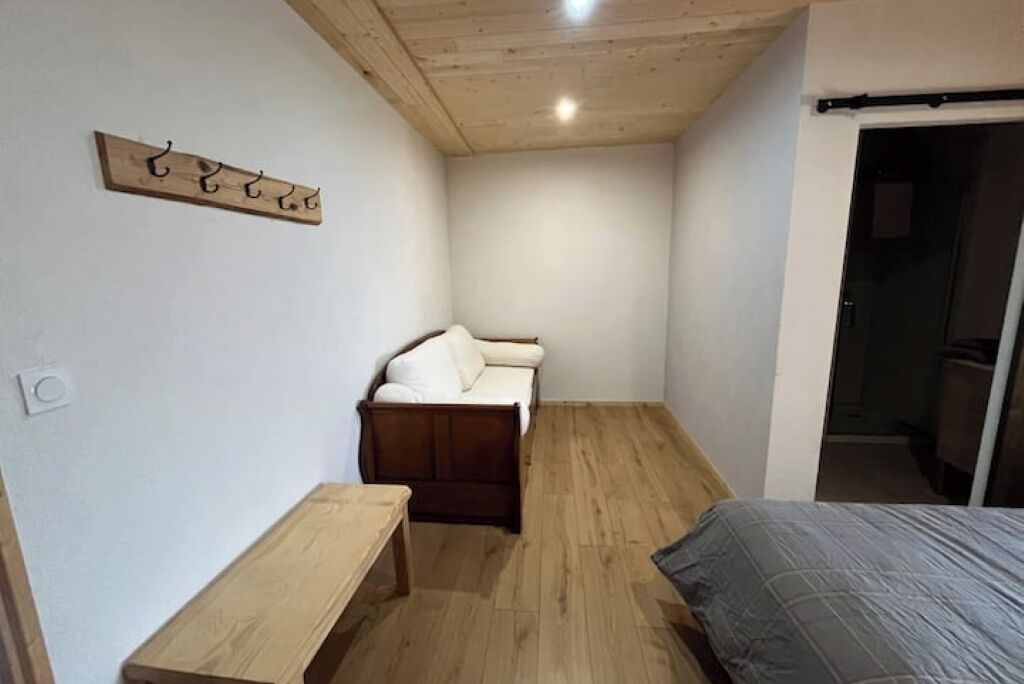 A small, narrow room with a wooden bench, a bed with a grey duvet, and a white sofa.