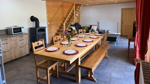 A dining area with a wooden table set for six, surrounded by chairs. A wooden bench is placed along one side of the table. A kitchen area with a microwave and stove is visible to the left. A staircase leads to an upper level.