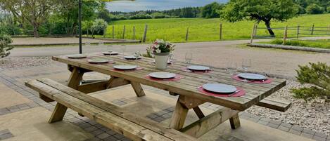 Une table de pique-nique en bois est installée à l’extérieur, sous un parasol jaune, et est entourée d’un chemin de gravier et de champs verdoyants.