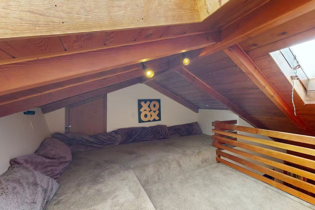 Un dormitorio tipo loft con techo inclinado, vigas de madera, una cama con manta gris, una obra de arte colgada en la pared y una barandilla de madera.