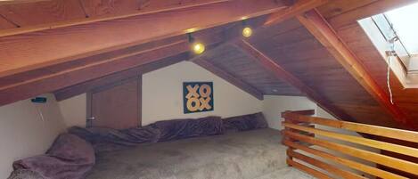 Un dormitorio tipo loft con techo inclinado, vigas de madera, una cama con manta gris, una obra de arte colgada en la pared y una barandilla de madera.