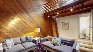 Une pièce chaleureuse de style loft, avec un plafond en pente, des poutres en bois et une échelle intégrée. On y trouve deux canapés gris ornés de coussins à motifs, une petite table avec une lampe et un tableau encadré accroché au mur.
