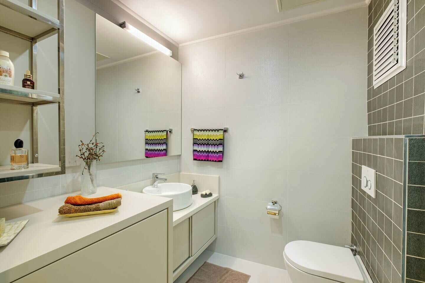 A modern bathroom with a white sink, mirror, and toilet. There are colorful towels hanging on the wall and a small shelf with toiletries.