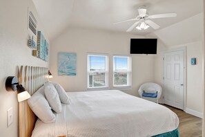 A bedroom with a large bed, two windows, a ceiling fan, a mounted TV, and a chair by the door.