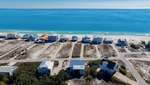 Vue aérienne d’un quartier résidentiel situé sur la côte, où les maisons sont alignées le long de la plage, avec des routes de sable et une vue dégagée sur l’océan bleu.