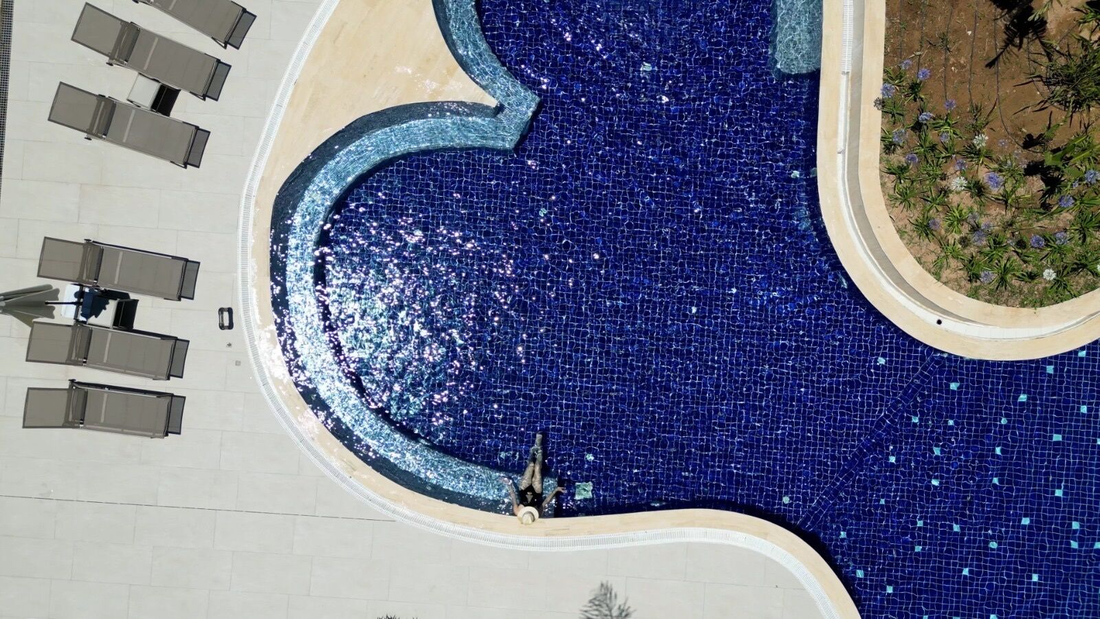 Ein Luftbild eines Schwimmbeckens mit gefliestem Rand, umgeben von einer gepflasterten Fläche mit Sitzgelegenheiten und einem kleinen Garten.