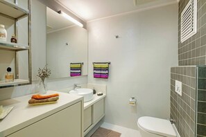 Een moderne badkamer met een wit wastafel, spiegel en toilet. Kleurrijke handdoeken hangen aan de muur en er is een klein plankje met toiletartikelen.