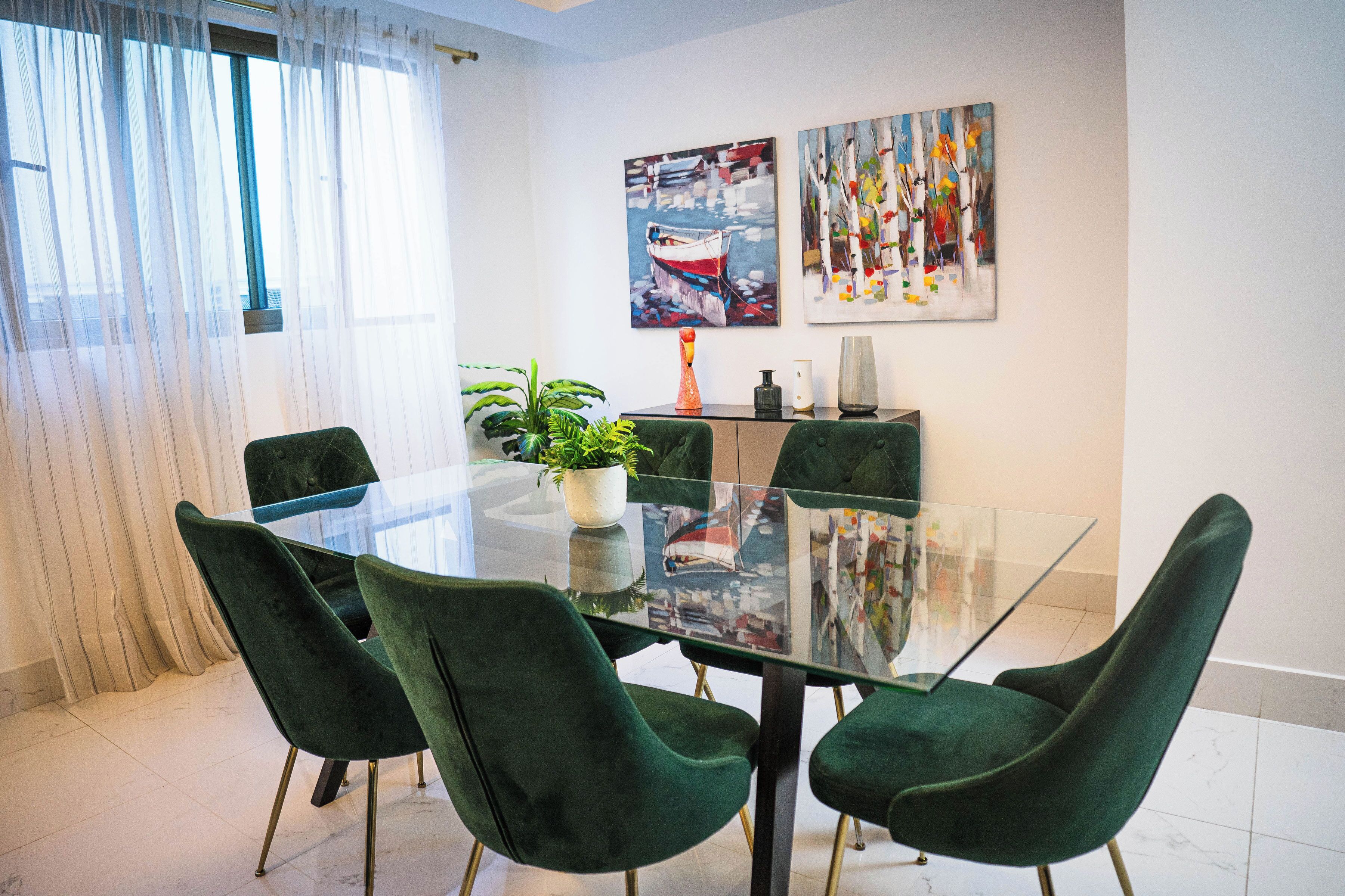 A dining area with a glass table, green upholstered chairs, a potted plant, and artwork on the wall.
