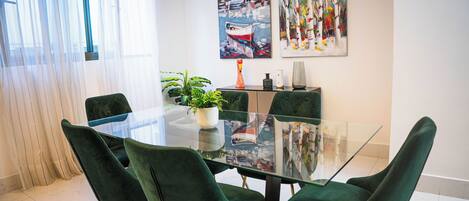 A dining area with a glass table, green upholstered chairs, a potted plant, and artwork on the wall.
