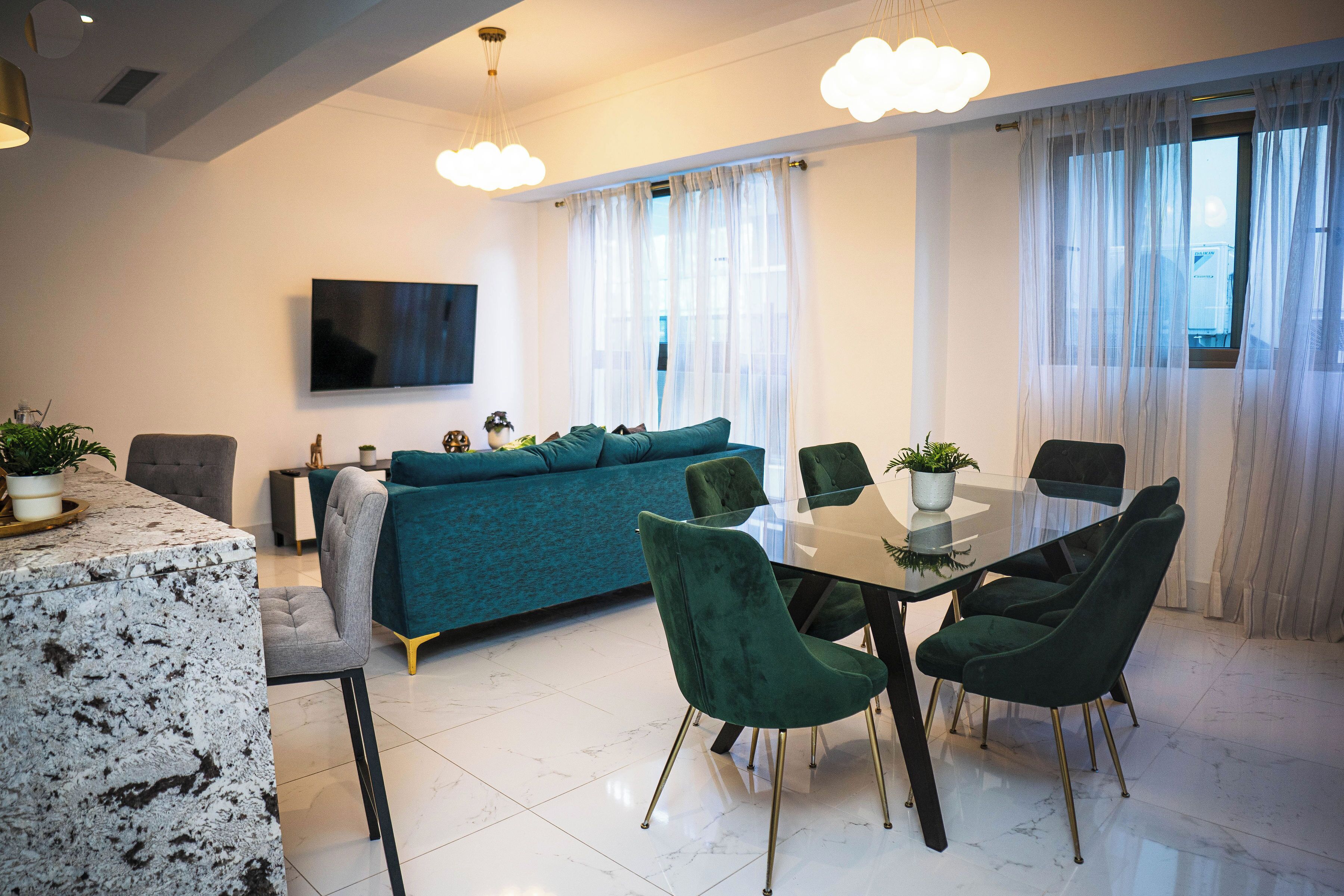 Una sala de estar moderna con un sofá color turquesa, una mesa de comedor de vidrio y sillas verdes. Un televisor de pantalla plana está montado en la pared.