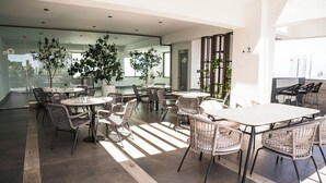 A modern indoor dining area with multiple tables and wicker chairs, surrounded by large potted plants and glass walls. - Arpel 07 - Piantini - Santo Domingo (Santo Domingo)