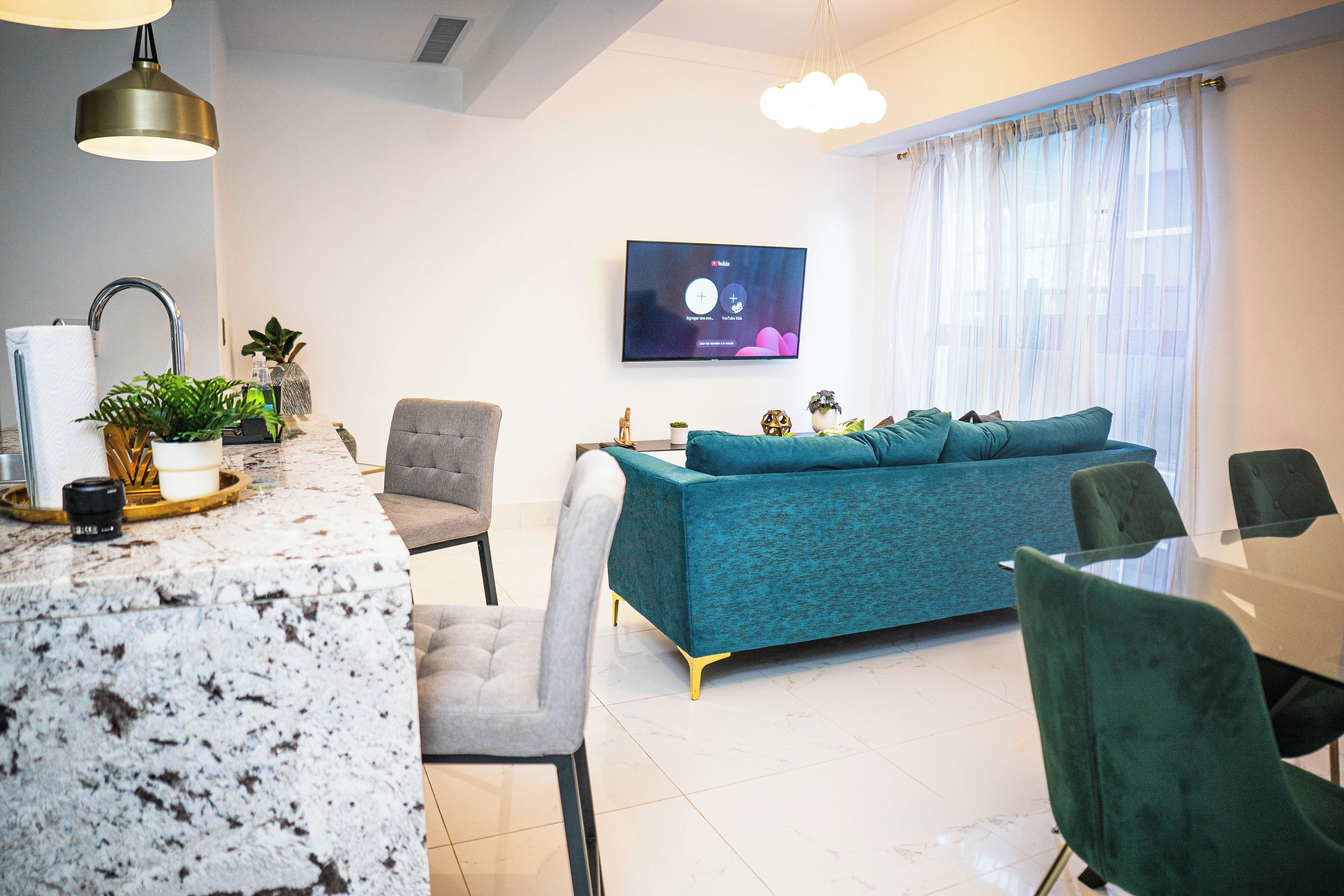 A modern living room with a teal sofa, a marble coffee table, and a flat-screen TV mounted on the wall.