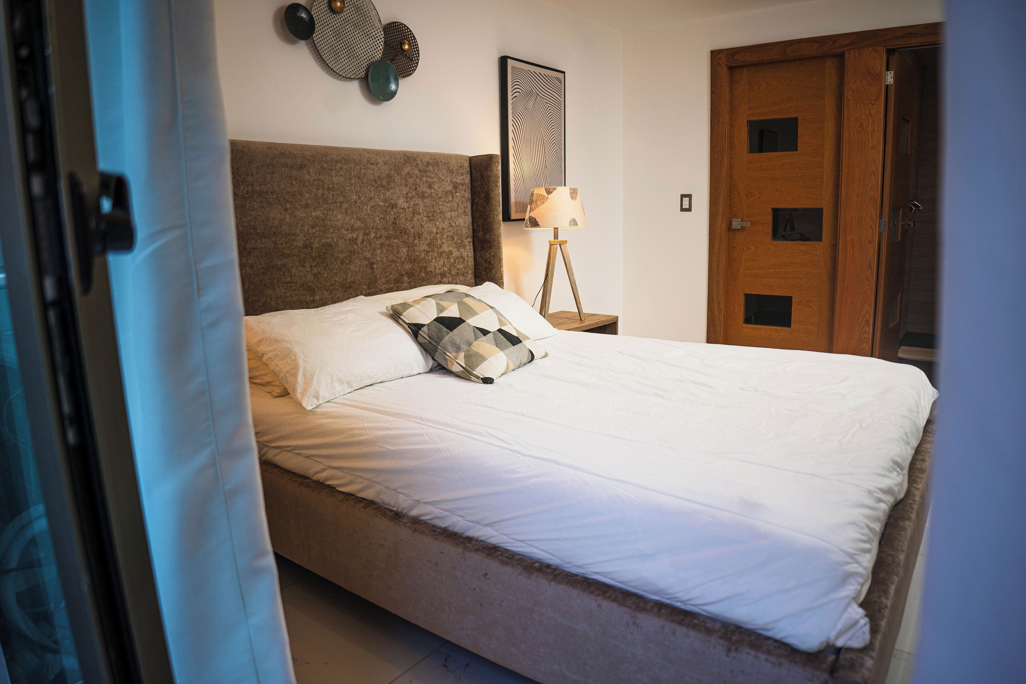 A neatly made bed with white linens and a brown headboard, a bedside lamp, and a wooden door with a small window.