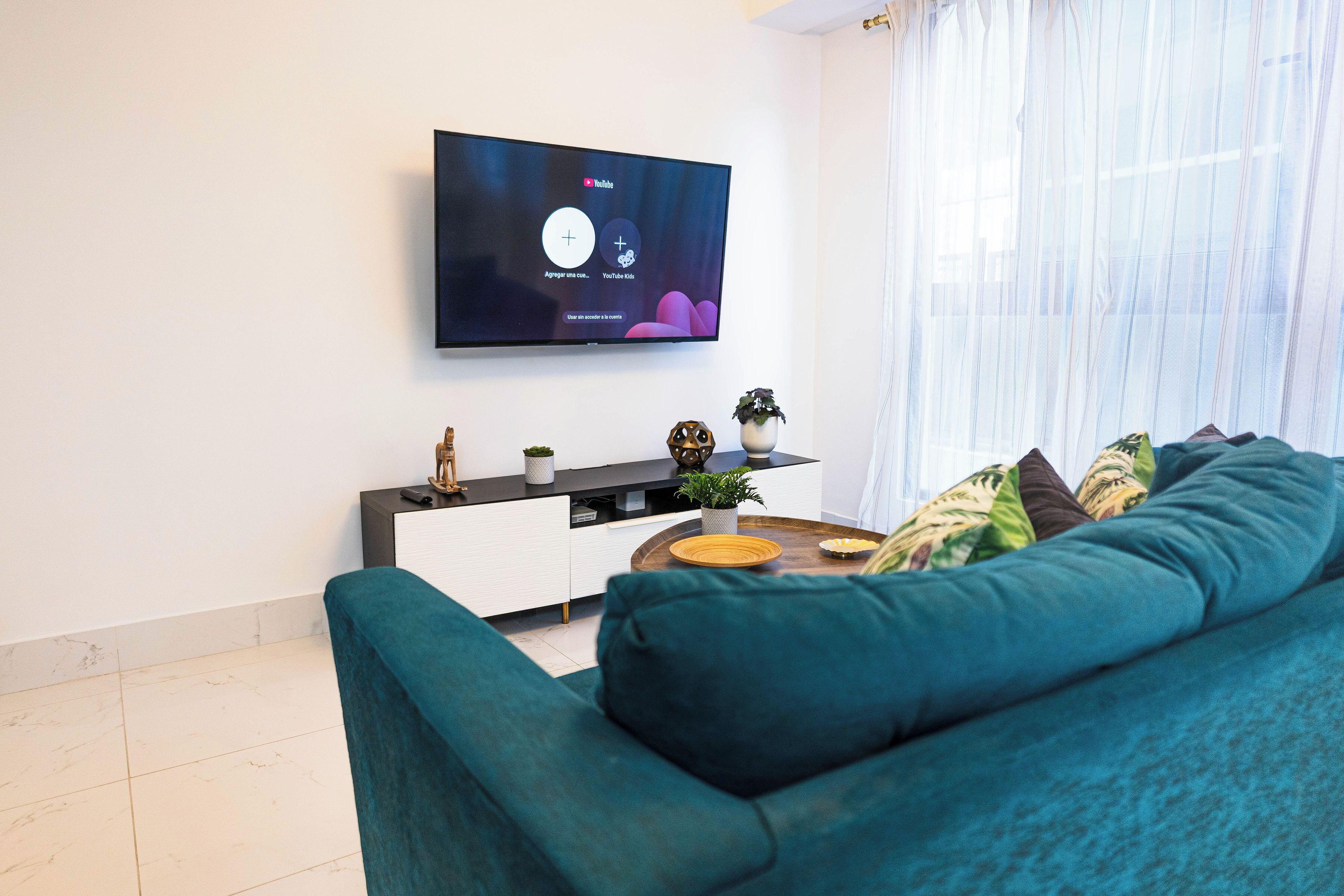 Un salon moderne avec un canapé de couleur turquoise, un téléviseur à écran plat fixé au mur, une petite table d’appoint agrémentée d’objets décoratifs et une grande fenêtre ornée de rideaux légers.