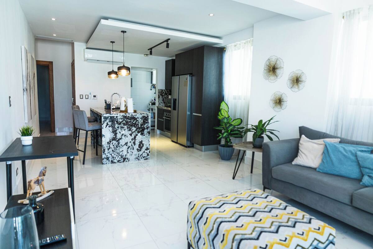 Una sala moderna con un sofá, un otomano estampado y un comedor con una isla de mármol.