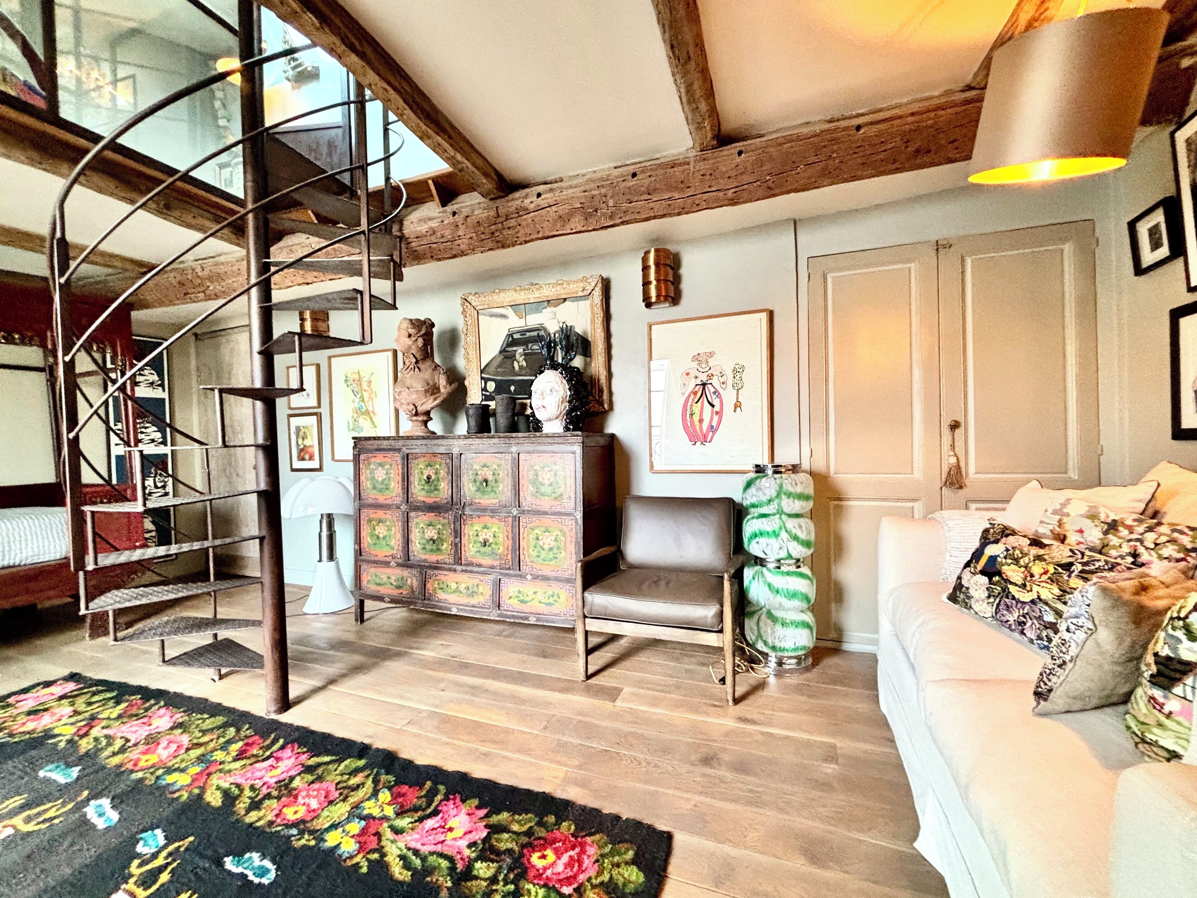 Una habitación amplia con una escalera en espiral, un armario de madera con objetos decorativos, un sofá con cojines estampados y una alfombra floral.