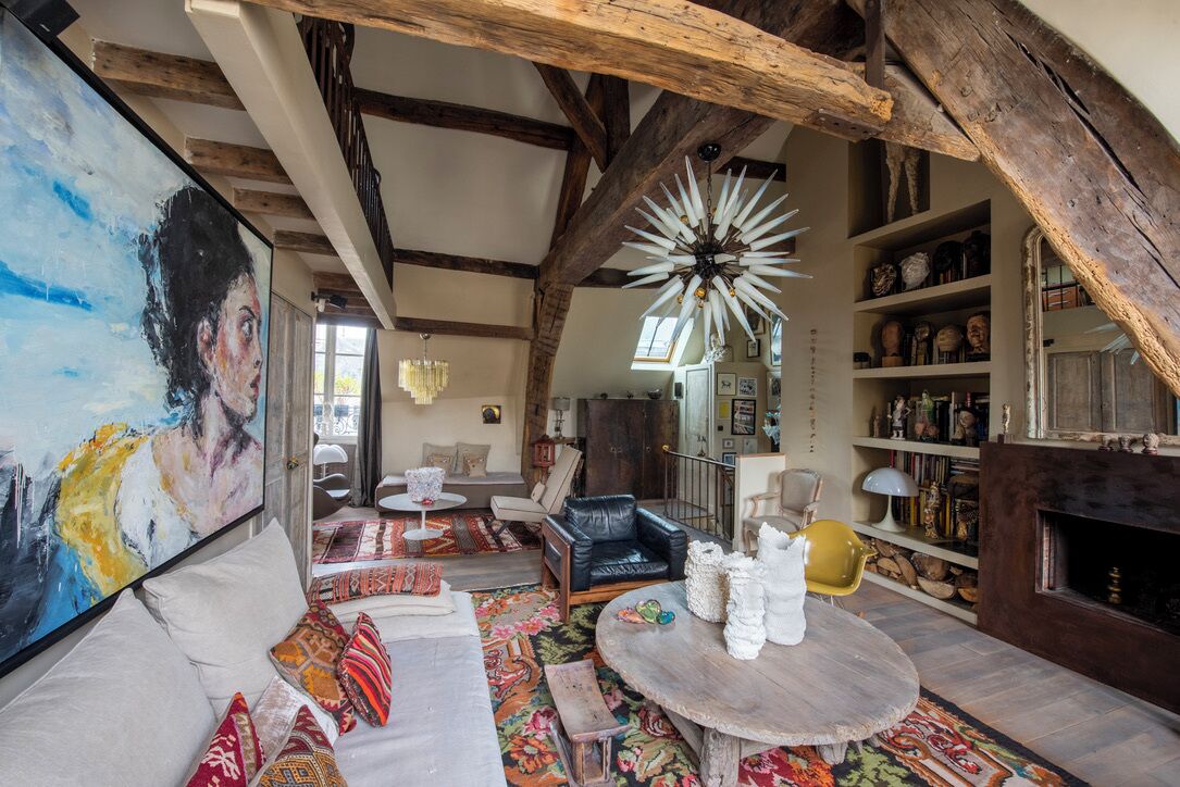 Un interior rústico con vigas de madera a la vista, una mesa redonda de madera y una estantería llena de libros y objetos decorativos.