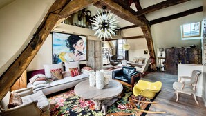 Un salón estilo loft con vigas de madera a la vista, un cuadro grande, una mesa de centro redonda y diversas opciones de asientos, incluyendo un sofá, sillas y un sillón mecedora.