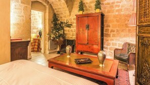 A cozy room with a stone wall, a wooden cabinet, a wicker chair, and a bed. - 18TH-CENTURY PALACE, BLEND OF OLD-WORLD GRANDEUR & MODERN COMFORT - LE MARAIS (Paris)