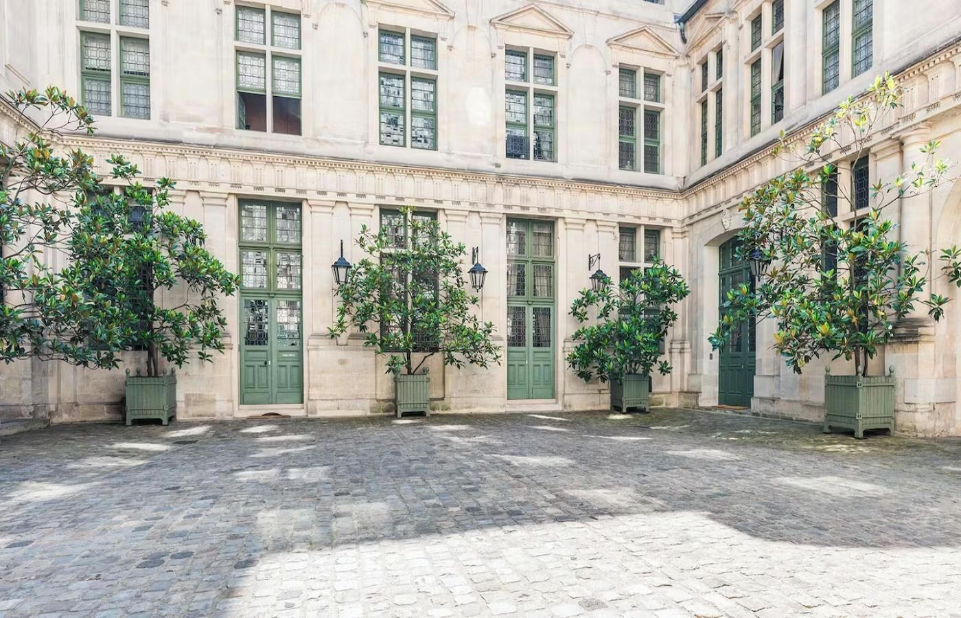 A courtyard with a cobblestone floor, surrounded by classical architecture with green doors and windows.