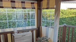 Una terraza de madera con un banco, una mesita con un florero y una ventana con vistas a la vegetación.