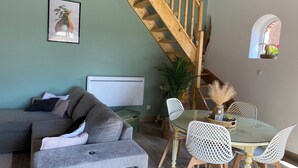 Ein gemütliches Wohnzimmer mit grauem Sofa, einem Glastisch mit weißen Stühlen, einer Holztreppe und einem kleinen Fenster mit Blick auf Grünflächen.