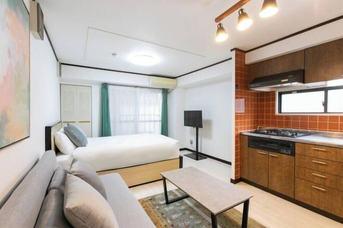 Habitación compacta con cama, sofá, mesita, kitchenette y ventana con cortinas.
