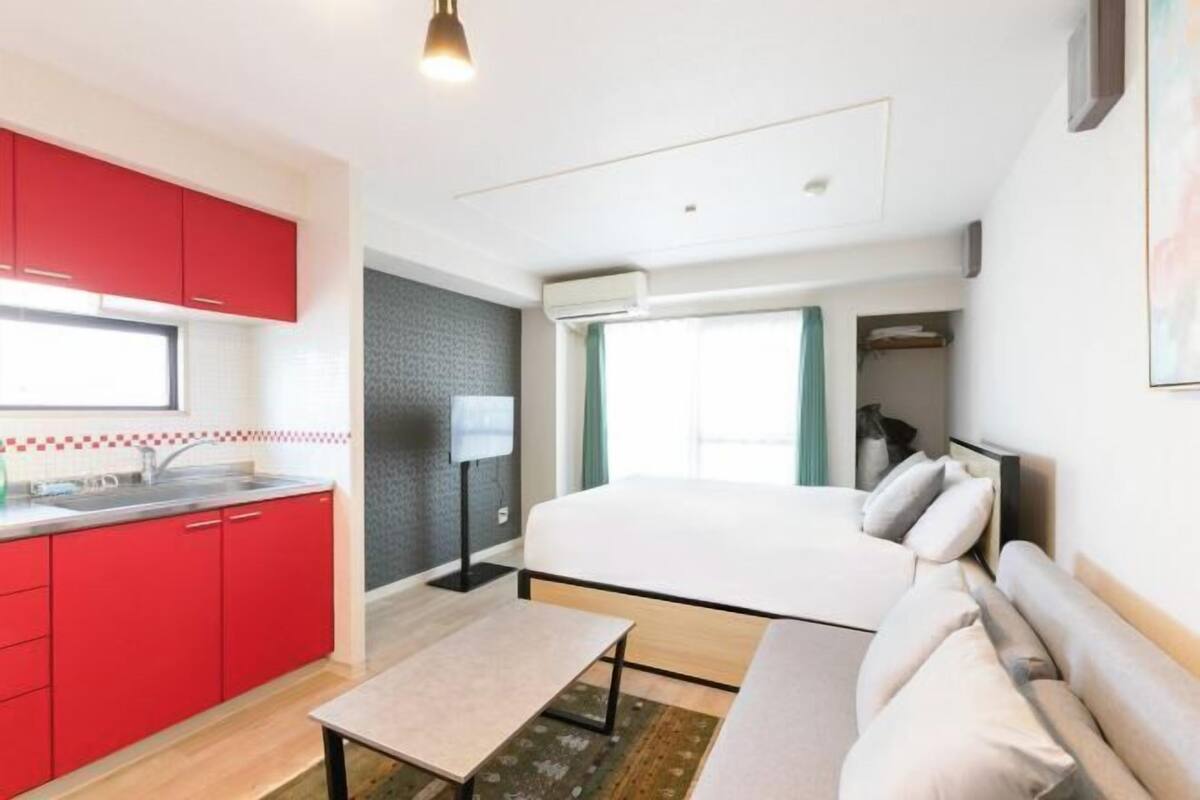 Habitación compacta con cama, una mesita, un sofá, una kitchenette con armarios rojos y una ventana con cortinas.