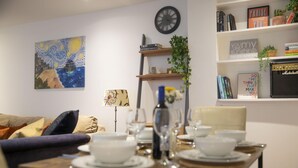 A dining area with a table set for two, a bookshelf with books and decorative items, a clock on the wall, and a painting above the couch. - Powis Patio Retreat By My Getaways (Brighton)