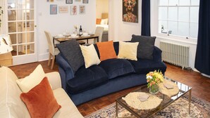 A living room with a dark blue sofa, a beige sofa, and a coffee table with a vase of flowers. There is a dining area with a table and chairs, and a window with curtains. - Powis Patio Retreat By My Getaways (Brighton)