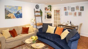A living room with a beige sofa, a blue sofa, a coffee table, and a patterned rug. There is a painting on the wall and a bookshelf with books and decorative items. - Powis Patio Retreat By My Getaways (Brighton)