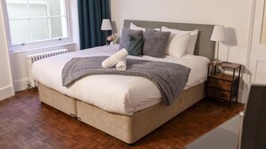 A neatly made bed with a grey blanket and pillows, a bedside table with a lamp, and a window with curtains. - Powis Patio Retreat By My Getaways (Brighton)
