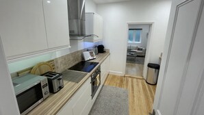 Et kompakt køkken med moderne hårde hvidevarer, et spiseområde med bord og stole, og udsigt til en stue med sofa og vindue.