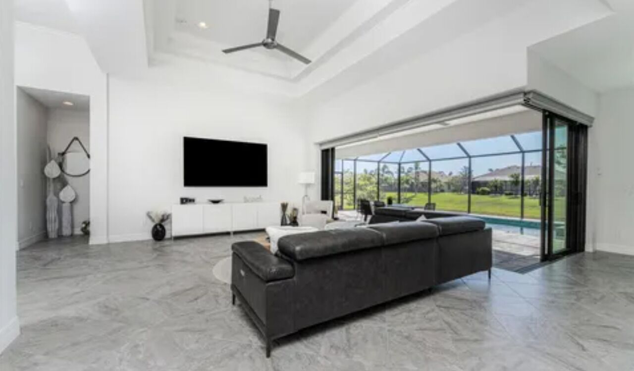 A spacious living room with a large gray sectional sofa, a flat-screen TV mounted on the wall, and a large sliding glass door leading to a pool area.