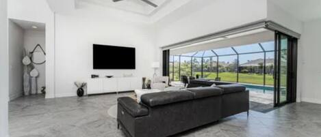 A spacious living room with a large gray sectional sofa, a flat-screen TV mounted on the wall, and a large sliding glass door leading to a pool area.