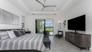 A modern bedroom with a large bed, a flat-screen TV, a ceiling fan, and a view through a glass door.