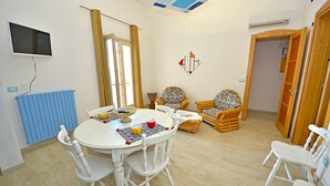 A dining area with a round table and white chairs, a blue radiator, and a television mounted on the wall. - Casa Mondrian - Camere con Bagno e Piscina Privata (Tiggiano)