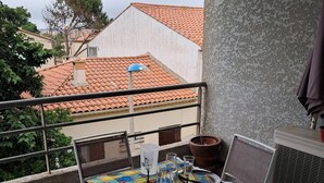 A balcony with a table set for two, chairs, a potted plant, and a window overlooking a building with a tiled roof. - Beautiful bright apartment on the seafront (Port-la-Nouvelle)