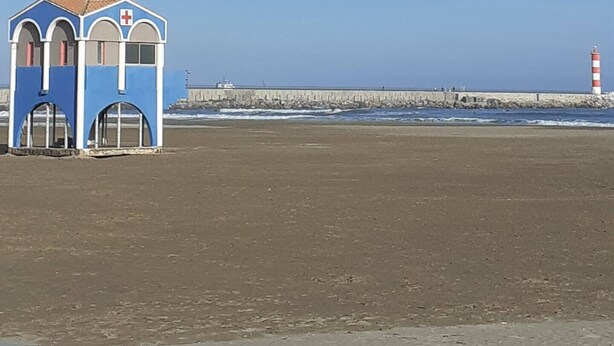 A beach with a blue building, a lighthouse, and a rocky breakwater. - Beautiful bright apartment on the seafront (Port-la-Nouvelle)
