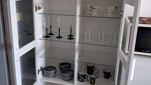 A white cabinet with multiple shelves containing various glassware, including wine glasses and tumblers, as well as black and white mugs. - Beautiful bright apartment on the seafront (Port-la-Nouvelle)